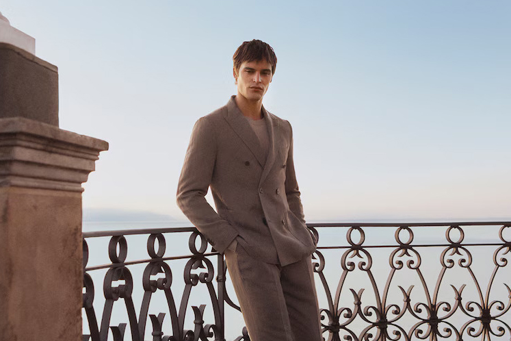 Man in brown suit on balcony overlooking water.